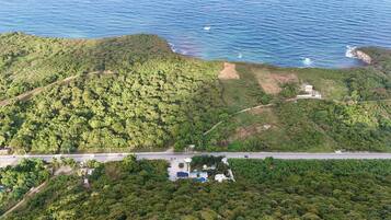 Vista aérea de una zona costera con abundante vegetación, una carretera y un pequeño edificio cerca de la orilla.