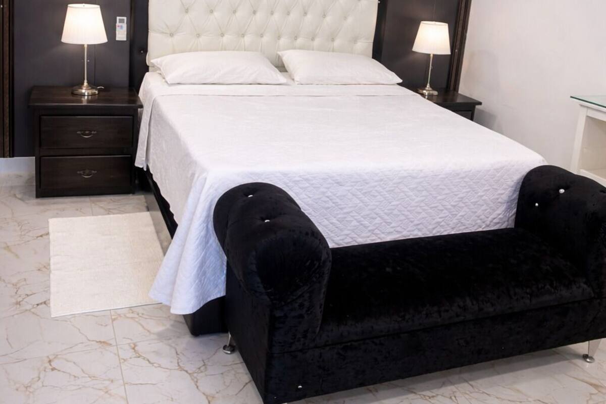 A bedroom with a large bed, two bedside tables with lamps, a dark headboard, a black bench, and marble flooring.