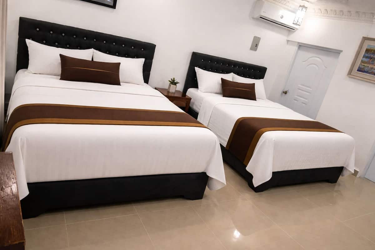 Two beds with black headboards and white bedding, a small bedside table with a plant, and a white door with a painting.