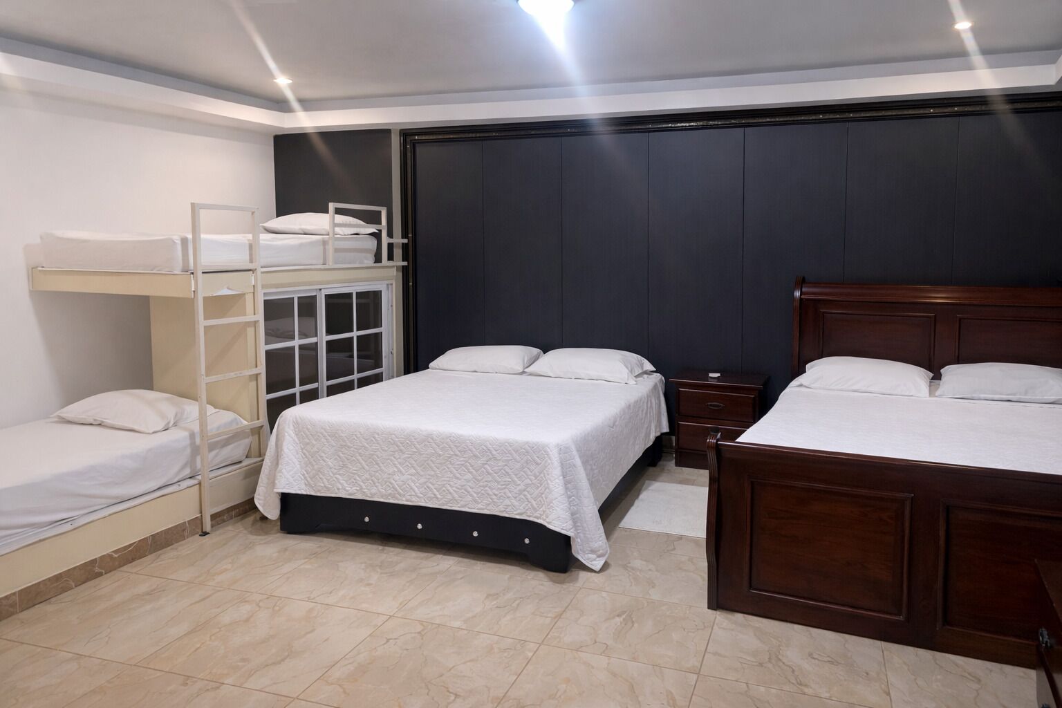 A bedroom with bunk beds, a single bed, a wooden headboard, a nightstand, and a dark wall.
