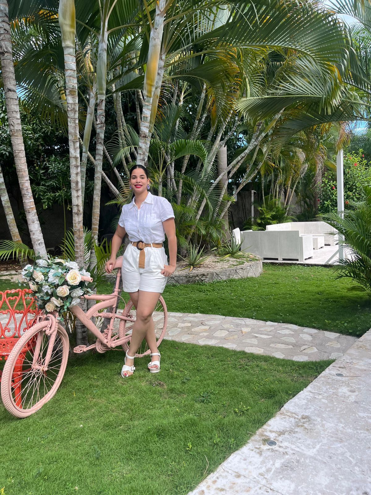 Eine Person steht neben einem pinkfarbenen Fahrrad mit einem Blumenkorb, in einem Garten mit Palmen und einer weißen Außenlounge.