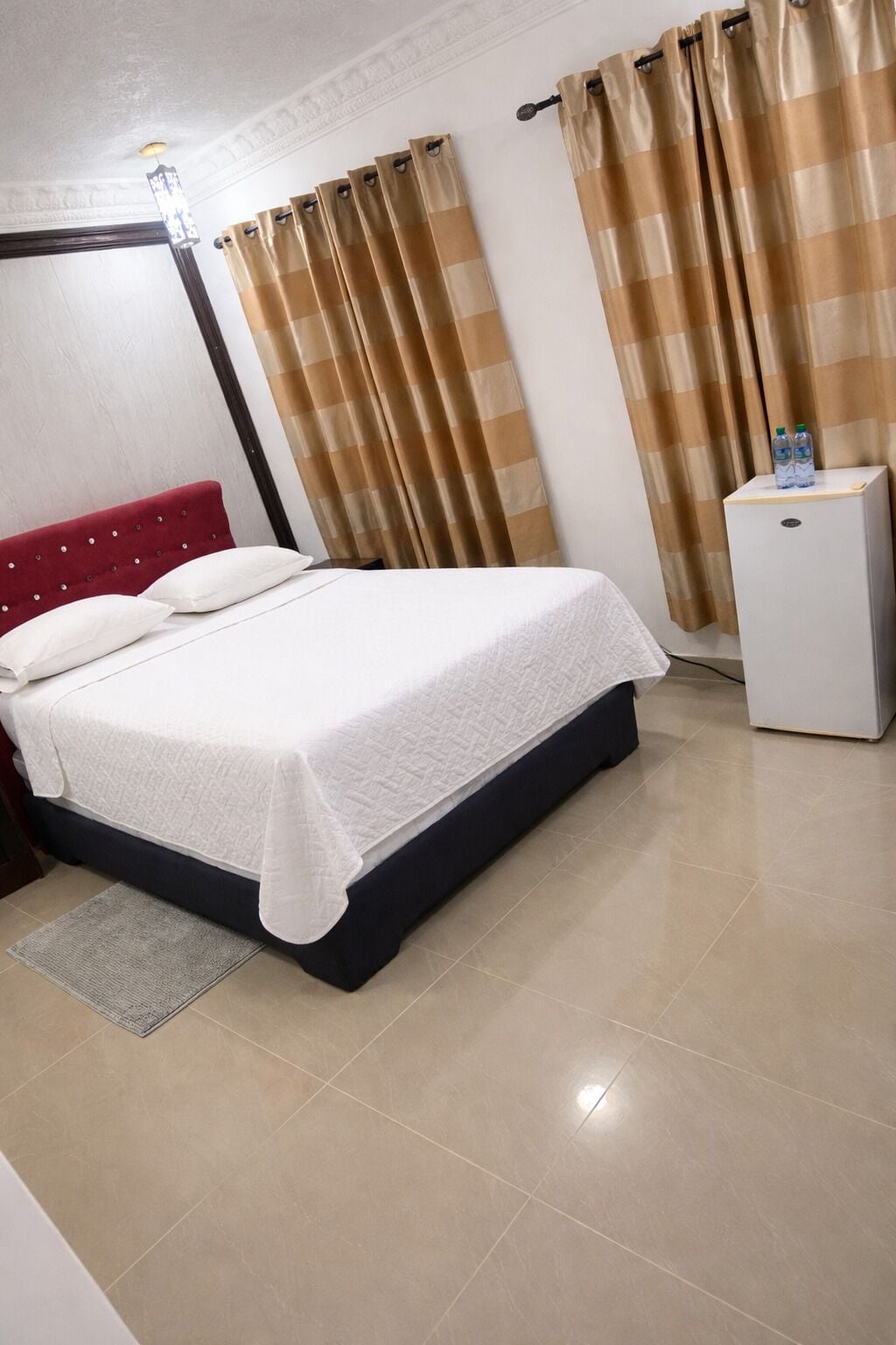 A hotel room with a bed, a small white refrigerator, and beige curtains.