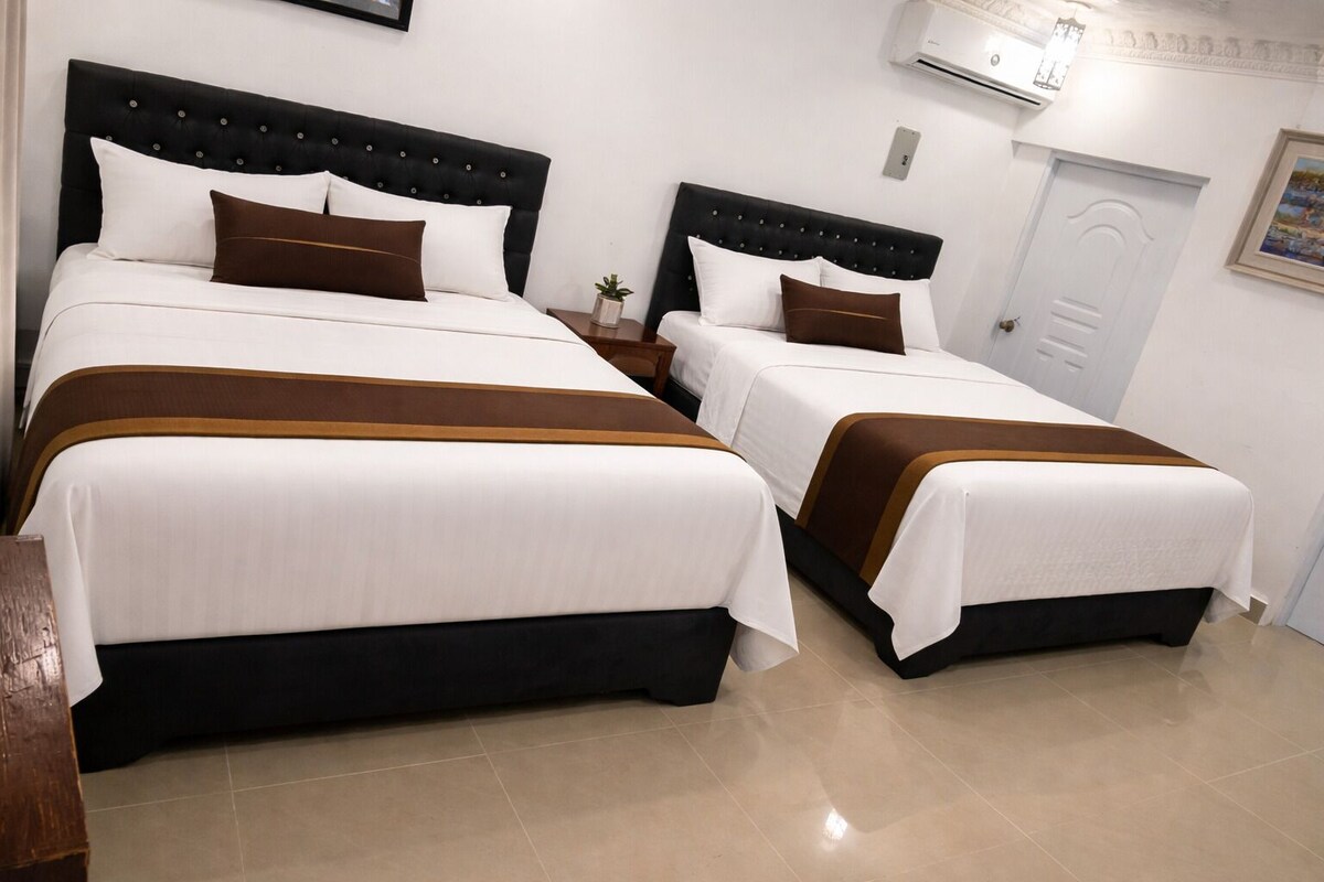 Two beds with black headboards and white bedding, a small bedside table with a plant, and a white door with a painting.