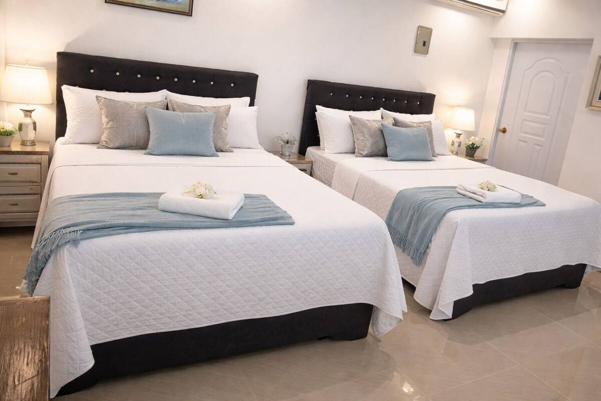 A hotel room with two beds, each with a dark headboard, white bedding, and a blue throw blanket. There are bedside tables with lamps and a door in the background.