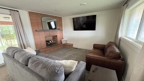 A living room with a grey sofa, a brown leather sofa, a small table, a TV mounted on the wall, and a brick fireplace.