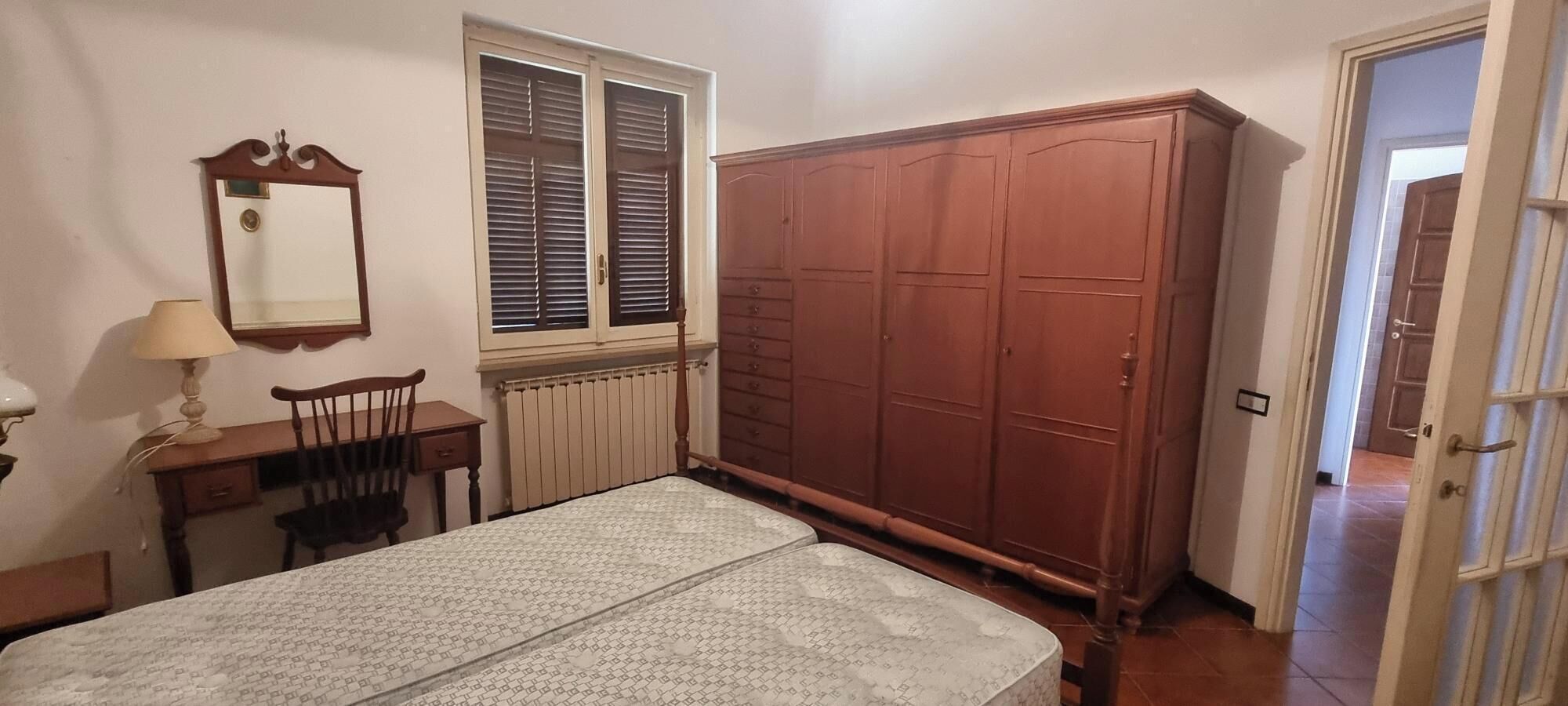 Ein Schlafzimmer mit einem Bett, einem Holzschrank, einem Schreibtisch mit Stuhl, einem Fenster mit Jalousien und einer Heizung.