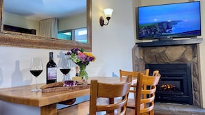 A dining area with a wooden table, two wine glasses, a bottle of wine, and a vase of flowers. A television is mounted above a stone fireplace.
