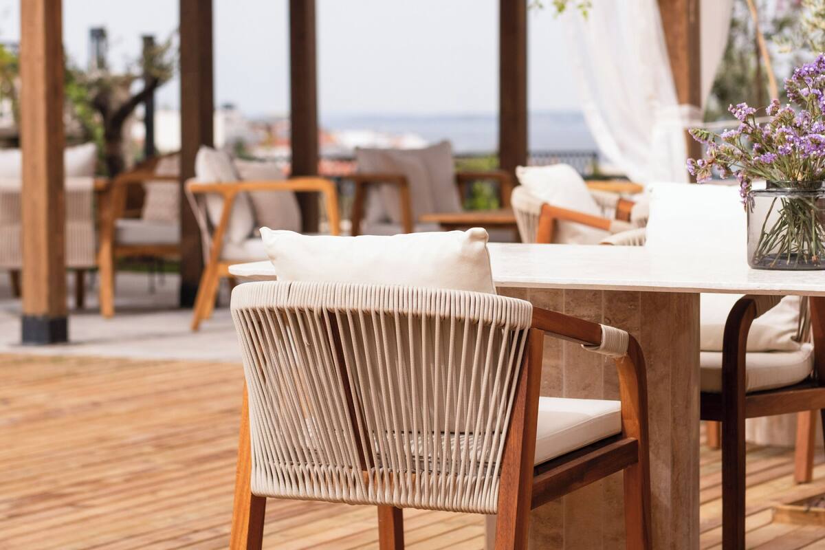 Una terraza de madera con una mesa y sillas, una pérgola con techo de tejas y vista al mar.