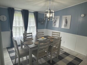 Uma sala de jantar com uma mesa posta para quatro pessoas, rodeada por cadeiras, um lustre pendurado e um relógio na parede.