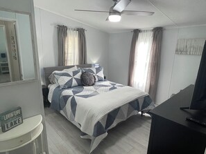 Quarto com cama, mesa de cabeceira, cômoda, ventoinha de teto e televisão.