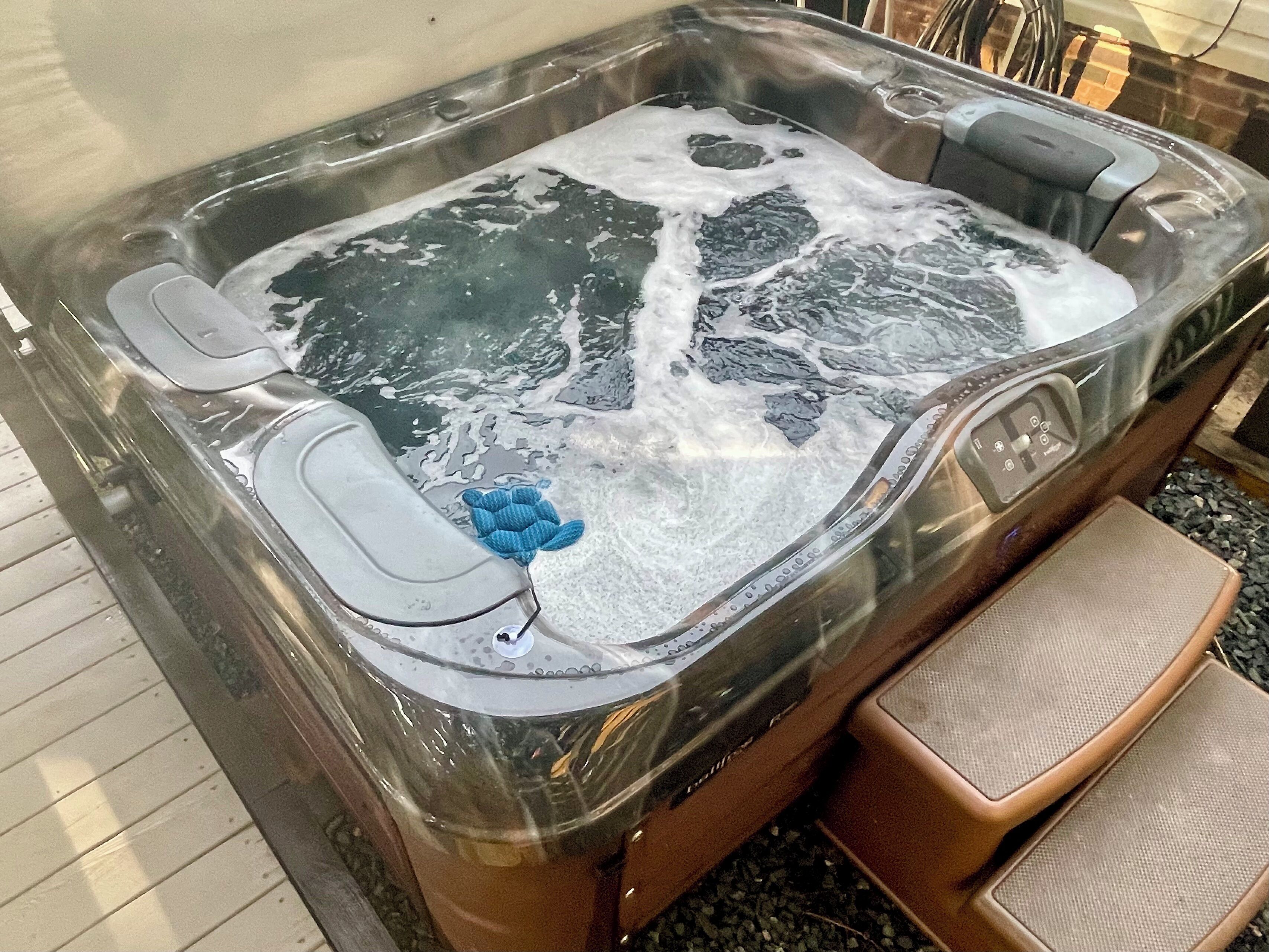 A hot tub with a clear glass enclosure, a control panel, and a pair of brown cushioned steps leading into it.