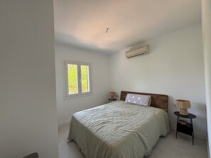 A bedroom with a bed, a window, an air conditioner, and a nightstand with a lamp. - Air-conditioned villa with private pool and large garden (Port d'Addaia)
