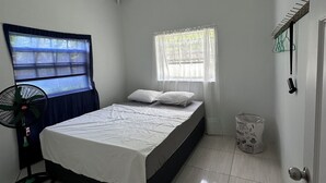 A bedroom with a bed, a window, a fan, and a trash bin.