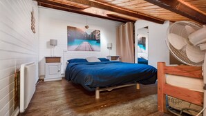 Un dormitorio con techo de madera, una cama con colcha azul, un radiador blanco y un ventanal con cortinas.