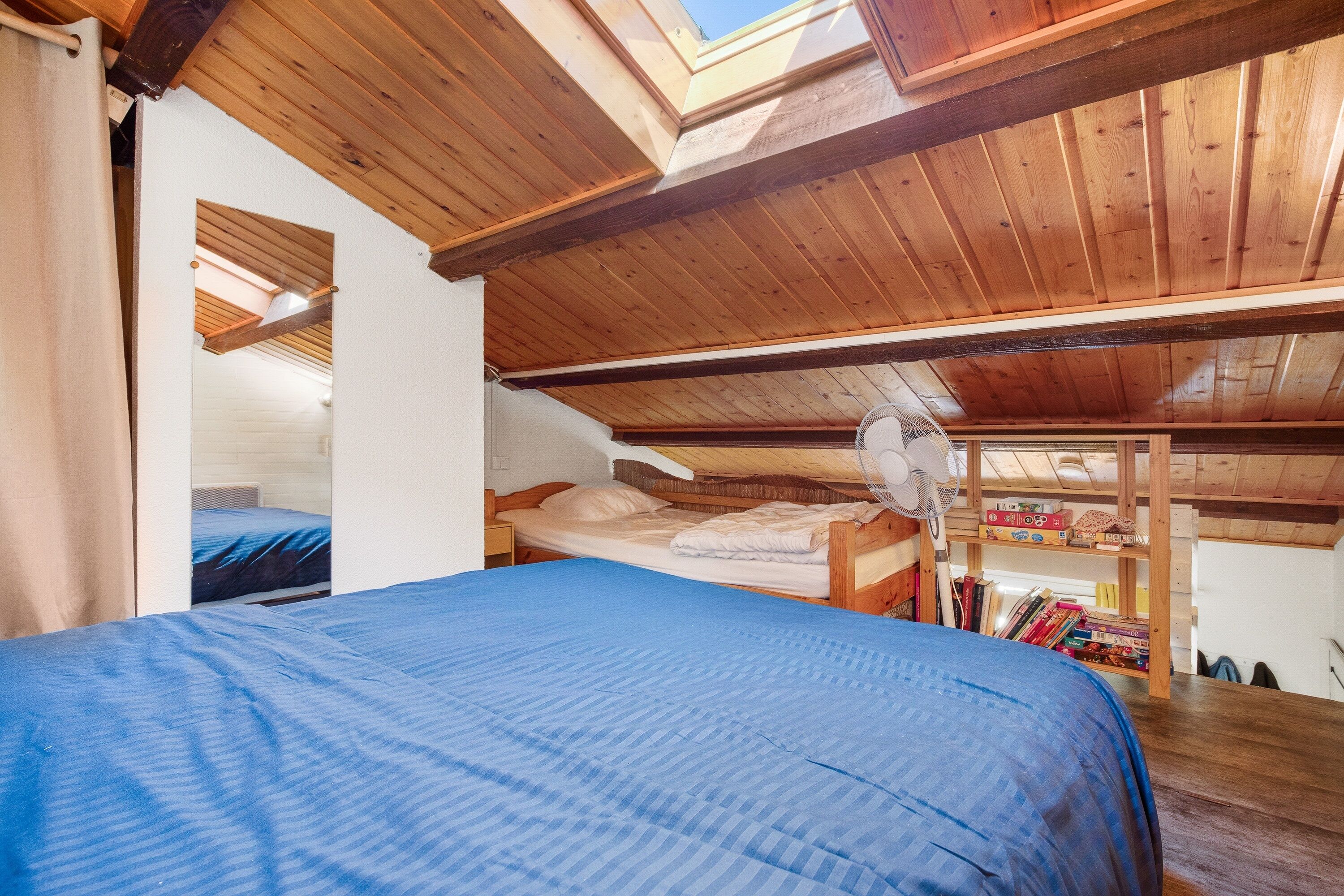 A room with a sloped ceiling, wooden beams, a skylight, a bed with a blue bedspread, a fan, and a shelf with books.