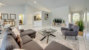 A spacious living room with a grey sofa set, a glass coffee table, and a grey armchair. There is a TV on a wooden stand and a large window providing natural light. - Heated Pool - Gulf Access WIDE canal with dock - BBQ - Large Backyard (Cape Coral)