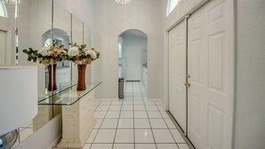 A hallway with a glass partition, white tiled floor, a large mirror, and a chandelier. - Heated Pool - Gulf Access WIDE canal with dock - BBQ - Large Backyard (Cape Coral)