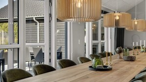 A long wooden dining table with chairs, large pendant lights, and a view of the outdoors through large windows. - 5 Star Holiday Home in Norre Nebel (Norre Nebel)