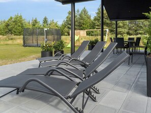 A covered outdoor seating area with lounge chairs and a table, surrounded by potted plants and a playground in the background. - 5 Star Holiday Home in Norre Nebel (Norre Nebel)
