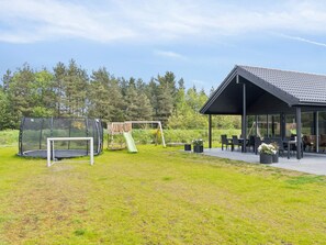 A covered outdoor seating area with tables and chairs, a playground with a slide, and a trampoline. - 5 Star Holiday Home in Norre Nebel (Norre Nebel)