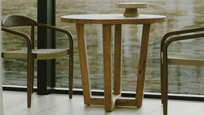 A modern interior with a round wooden table and two chairs, large windows with a view of a body of water and mountains.