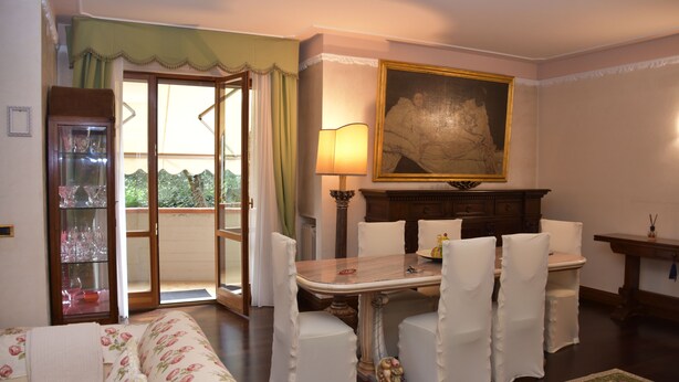 A dining area with a table set for two, white chairs, a lamp, and a painting on the wall.