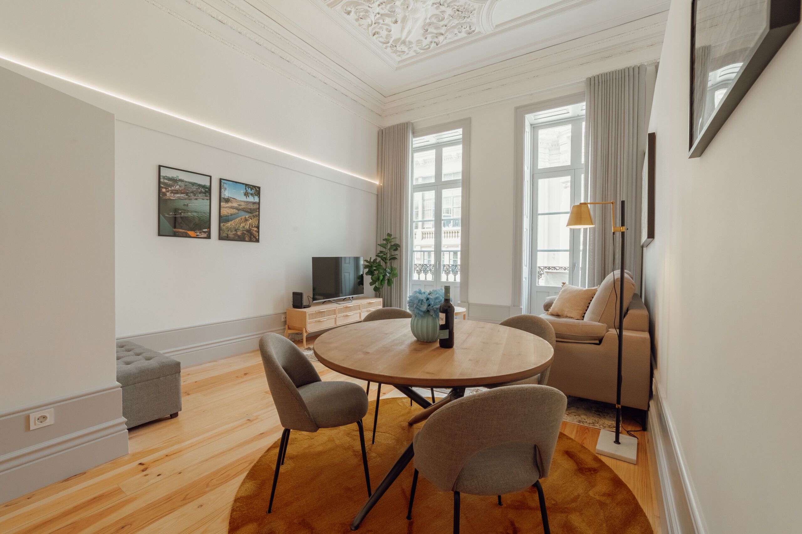 A modern living room with a round wooden table, grey upholstered chairs, a grey ottoman, a beige sofa, and a floor lamp.