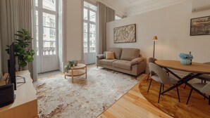 A living room with a beige sofa, a round wooden coffee table, and a dining table with chairs. There is a large window with curtains, a plant, and a painting on the wall. - Heart OF Porto Apartment A by Vacationy (Porto)