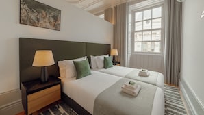 A hotel room with two beds, a nightstand with a lamp, a painting on the wall, and a window with curtains.
