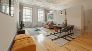A spacious living area with a sofa, a dining table with chairs, and a coffee table. There are large windows providing natural light and a view of the outside.