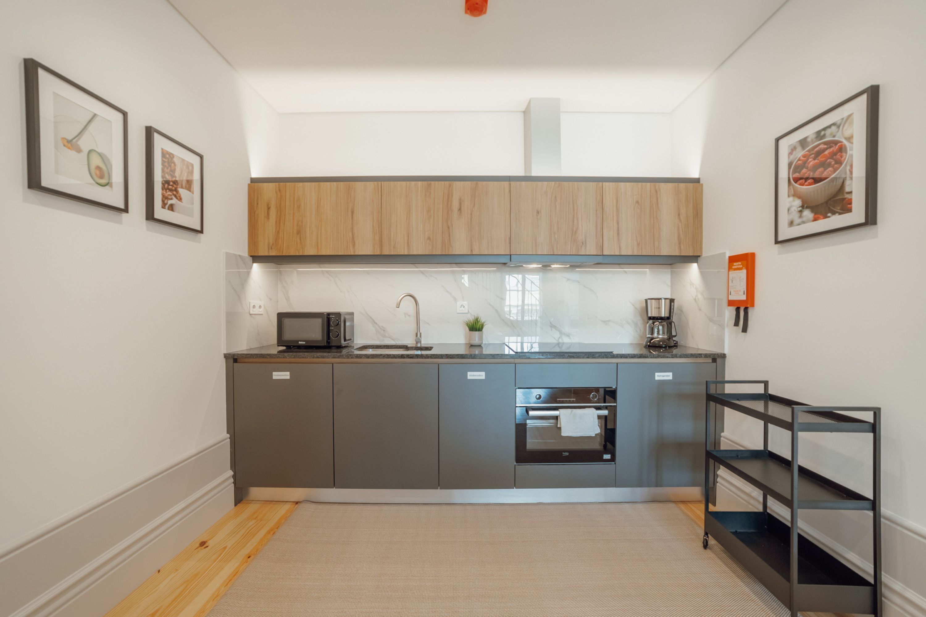 A modern kitchen with grey cabinets, a built-in oven, a microwave, and a coffee maker. There are two framed pictures on the wall and a small shelf unit.