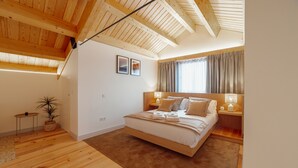 A bedroom with a wooden ceiling, a bed with beige bedding, a small table with a plant, and a window with curtains.