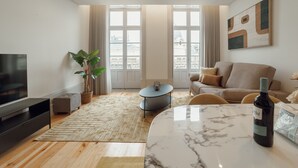 A modern living room with a TV, a marble table, a sofa, a potted plant, and a view of buildings through large windows.