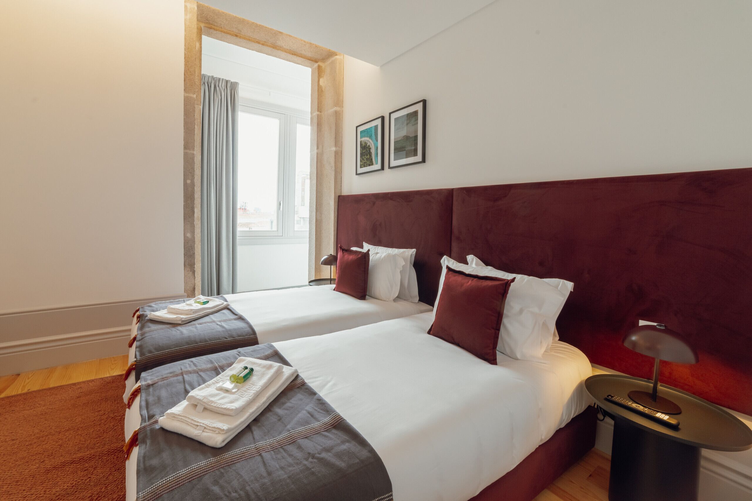 A hotel room with two beds, a large window with curtains, a red headboard, and a bedside table with a lamp.