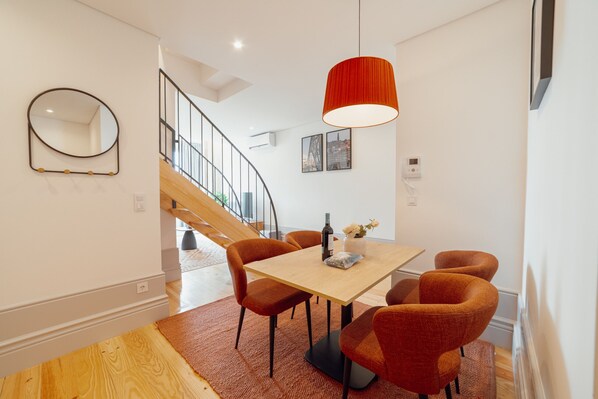 Un comedor moderno con una mesa de madera, sillas naranjas, una lámpara colgante roja y una escalera al fondo.