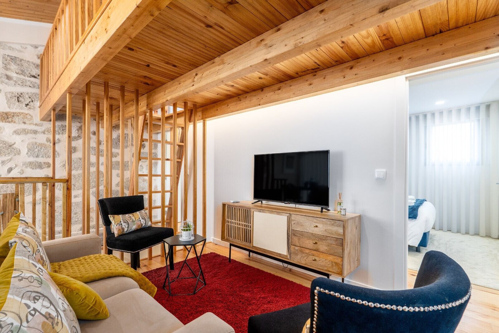 A modern living room with a wooden ceiling, a flat-screen TV, a sofa, a chair, and a red rug.