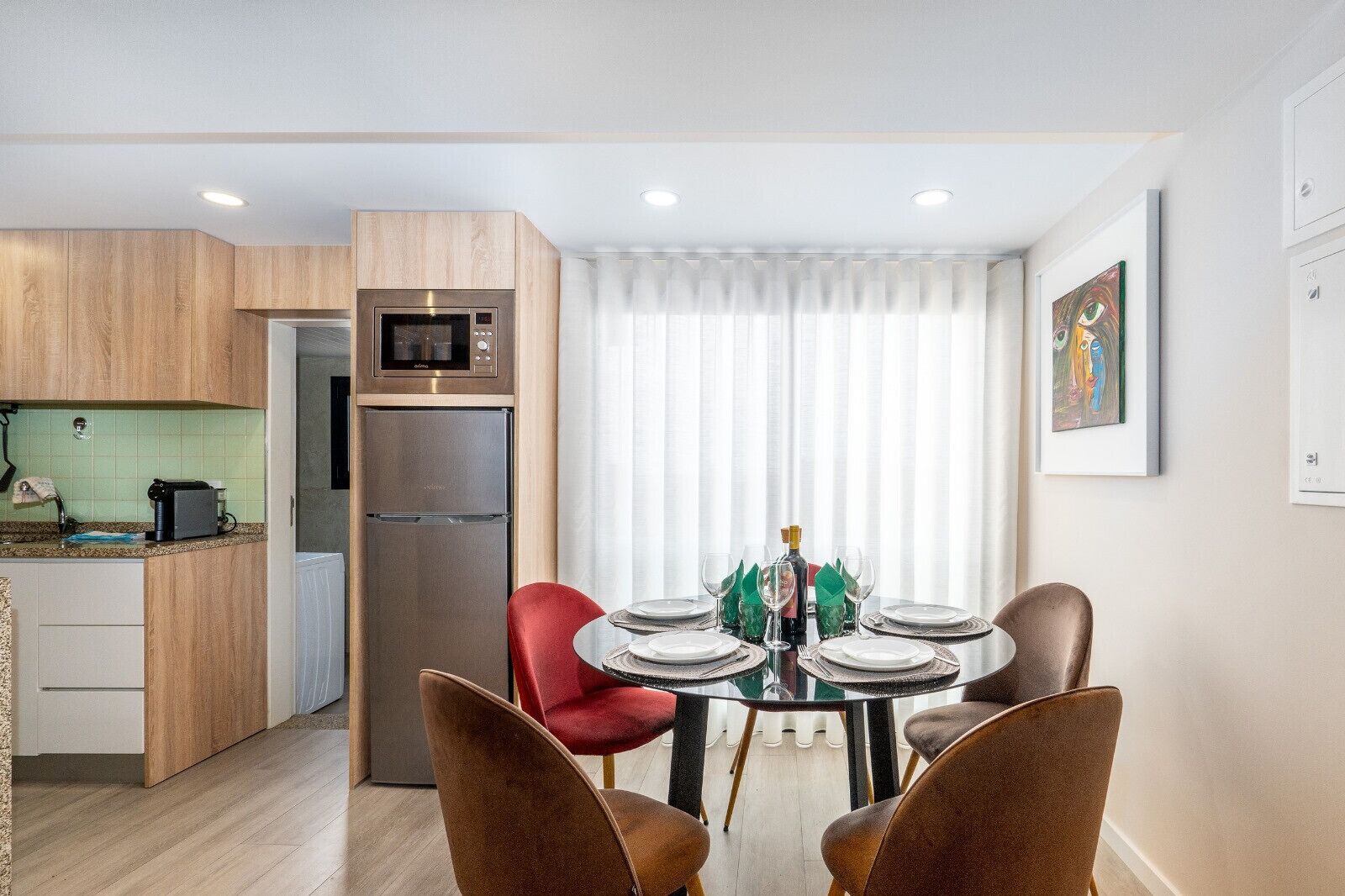 A modern kitchen with a built-in microwave and refrigerator, a dining area with a round table set for four, and a large window with sheer curtains.