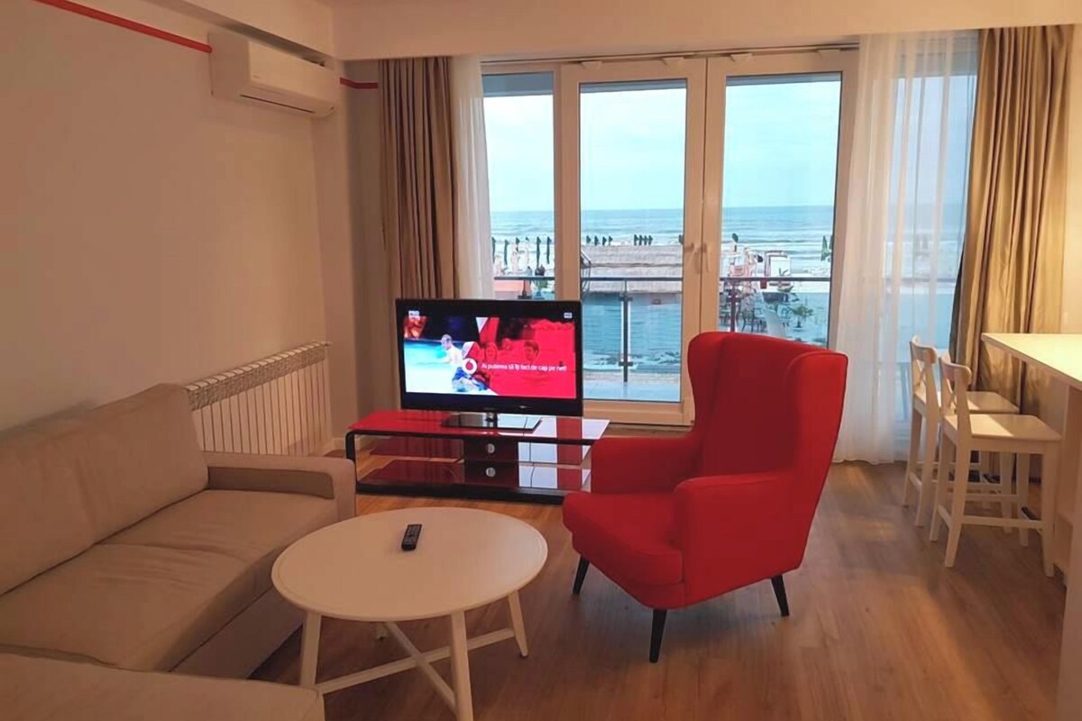 Un salon avec un canapé, un fauteuil rouge, une petite table ronde et un téléviseur diffusant un événement sportif.