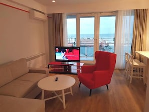 A living room with a sofa, a red armchair, a small round table, and a TV displaying a sports event.