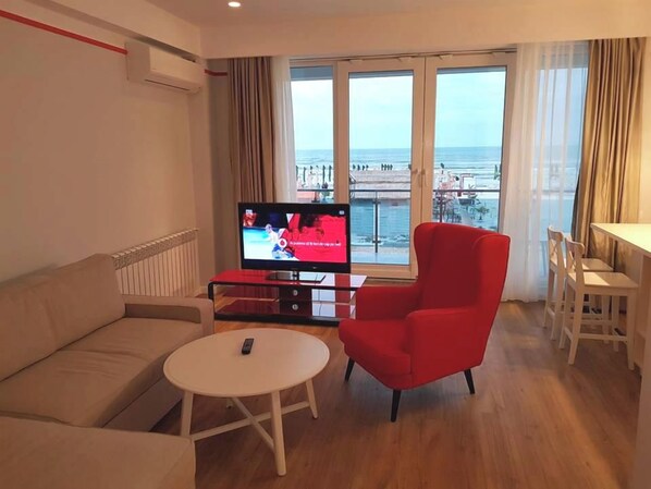 Un salon avec un canapé, un fauteuil rouge, une petite table ronde et un téléviseur diffusant un événement sportif.