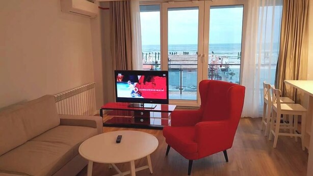 A living room with a sofa, a red armchair, a small round table, and a TV displaying a sports event. - Wave 1 Promenada Apartments (Navodari)