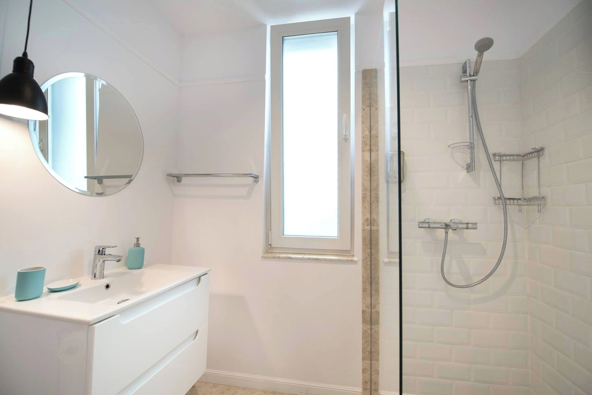 A bathroom with a white vanity, a round mirror, a window, and a shower with a handheld showerhead.