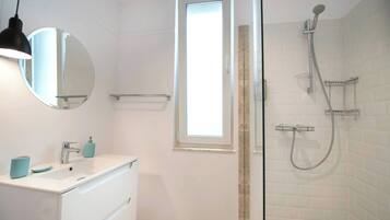 A bathroom with a white vanity, a round mirror, a window, and a shower with a handheld showerhead.