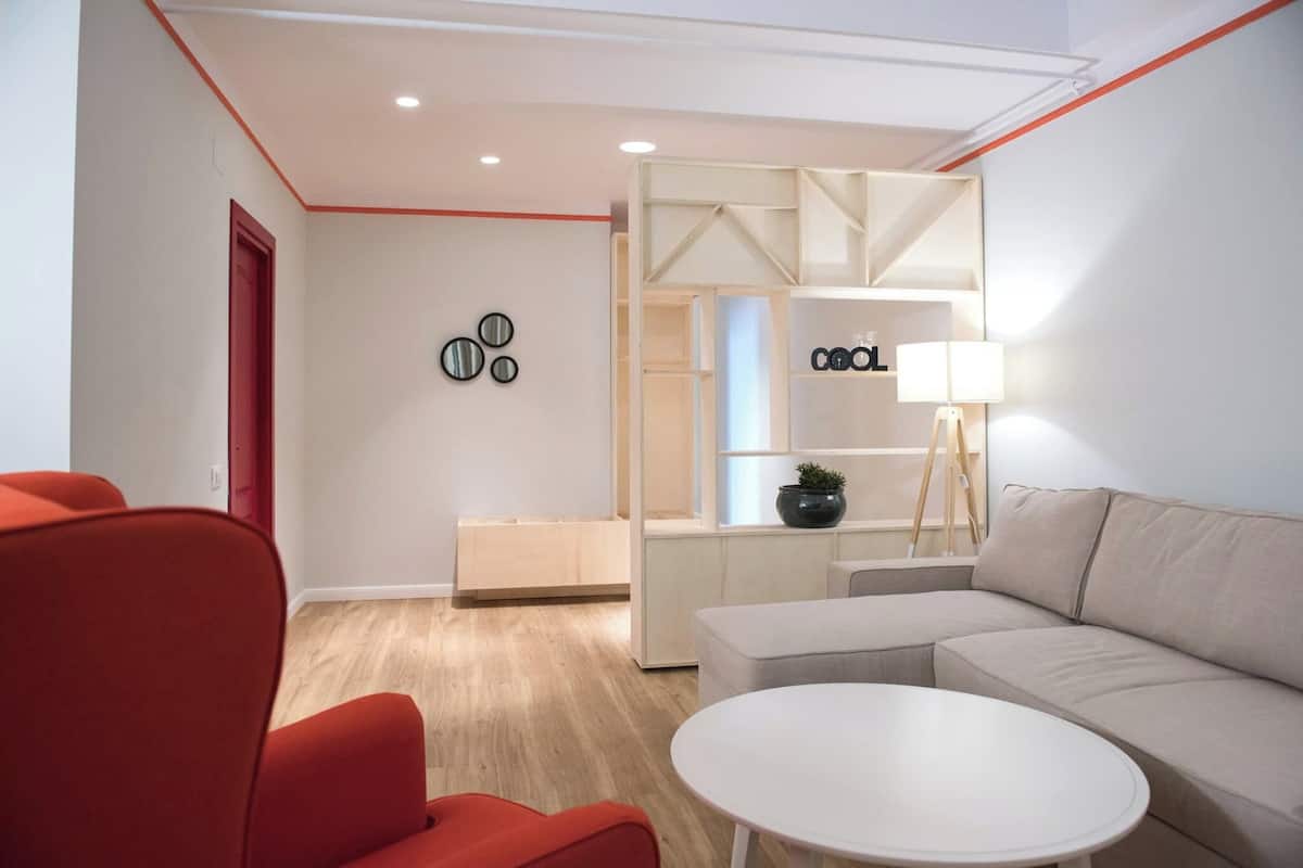 Un salon moderne avec un canapé, des fauteuils rouges, une table basse blanche et une étagère minimaliste agrémentée d’une lampe.