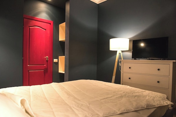 A bedroom with a bed, a red door, a dresser, and a lamp.