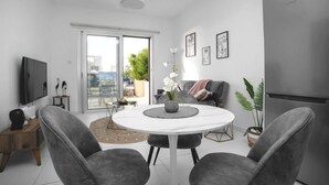 En moderne stue med et rundt hvidt bord, grå polstrede stole, et fladskærms-tv og en skydedør til en balkon.