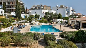 En swimmingpool omgivet af liggestole og parasoller, med bygninger i baggrunden.