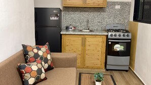 A compact living space with a kitchenette, a sofa with patterned cushions, and a small table with a plant. - Residencial Fe y Esperanza (Pedernales)