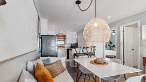 A modern living room with a sofa, dining table, and chairs. There is a kitchen in the background with cabinets and a refrigerator. - Shell Shocked - Short, Breezy Walk to the Beach (Carolina Beach)