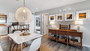 A modern kitchen with white cabinetry, a dining table with chairs, a hanging light fixture, and a wooden sideboard with baskets. - Shell Shocked - Short, Breezy Walk to the Beach (Carolina Beach)